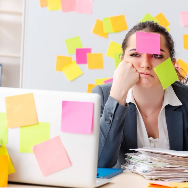 woman who looks overwhelmed by the amount of work she has to complete. She looks discouraged and has pot-it notes on her face and everywhere around her.